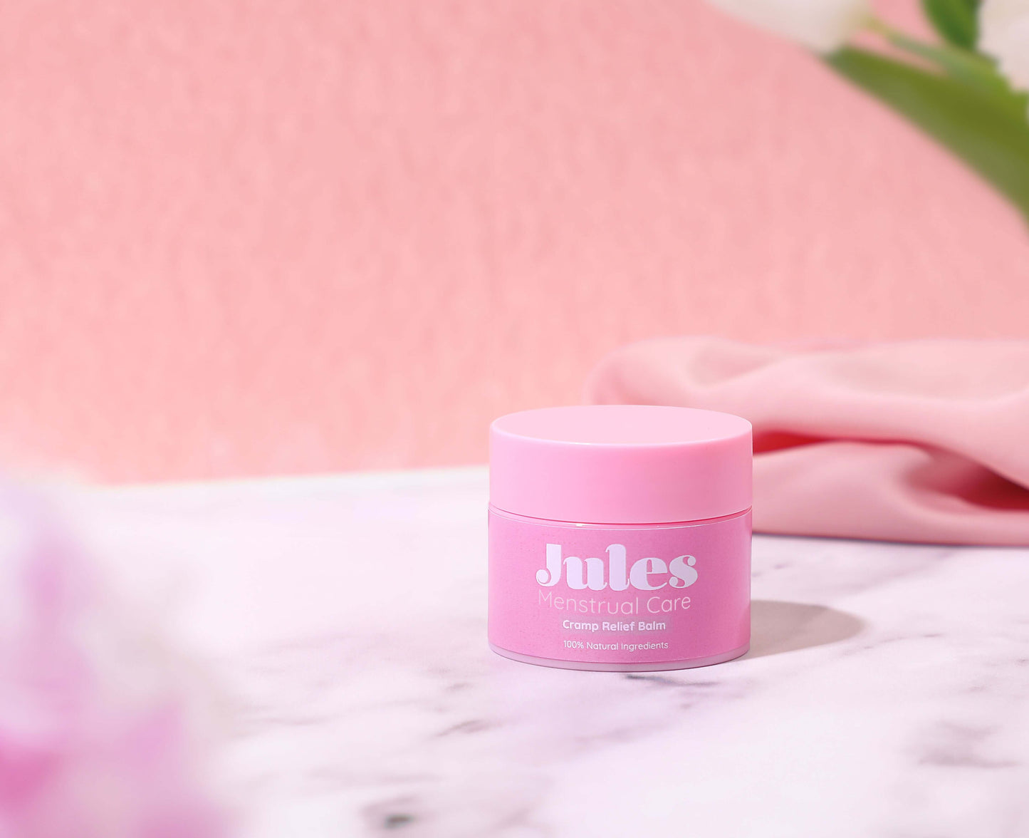 Cramp Relief Balm (30ml) in a cute pink jar surrounded by soft fabrics and flowers on a marble surface.